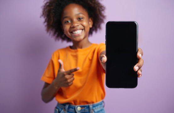 Happy African American kid girl holds new smartphone blank screen. Child points finger at mobile display, smiling widely. Youth promotes app or content on purple studio background.