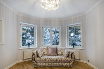 A cozy living room features a floral-patterned couch facing three windows. Outside, snow blankets the ground and trees, creating a peaceful winter scene