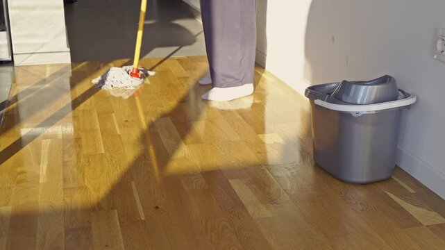 A person diligently mops a shiny wooden floor with a bucket, performing essential household chores. This scene highlights the effort of home maintenance and cleanliness in a bright, inviting space.