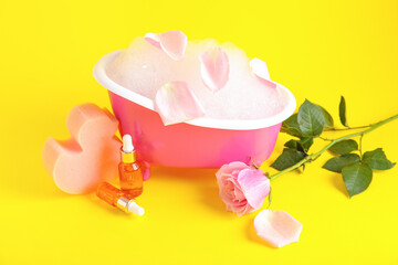 Small bathtub with foam, sponge, bottles of essential oil and rose flower on yellow background