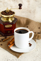 Cup of tasty coffee with cinnamon and grinder on light background