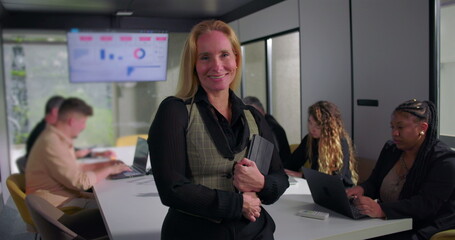 Smiling businesswoman standing confidently with tablet in hand during office meeting, supportive team working behind her in modern professional workplace setting