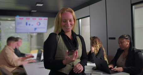Smiling businesswoman standing in office meeting room holding smartphone, diverse colleagues working on laptops with data charts on screen in background