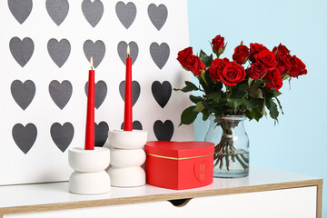 Bouquet of beautiful roses, candles and gift box on commode near blue wall at home decorated for Valentine's Day celebration
