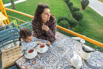 A mother and daughter enjoying tea together on a cafe balcony with a scenic green view, spending quiet family time outdoors and relaxing in a peaceful morning atmosphere.