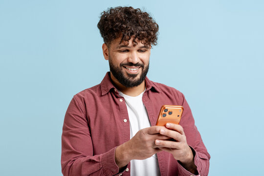 Smiling young man with curly hair texting on smartphone against pastel blue background