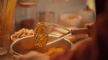 Hands are kneading cooked pumpkin with a masher to create a smooth texture for a creamy pumpkin soup. This cooking activity takes place in a warm kitchen, creating a comforting atmosphere
