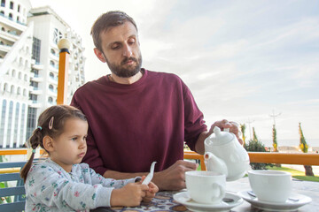 Father and daughter drinking tea together at an outdoor caf , enjoying peaceful family time on a balcony with a scenic view during a calm morning.