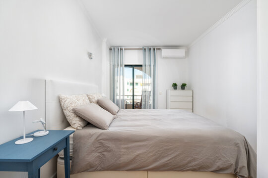 This bedroom features a large bed with soft linens, a small blue nightstand, and light curtains letting in natural light. A minimalist design enhances relaxation