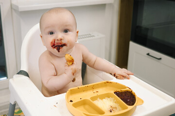 Toddler covered in food sits in a high chair and enthusiastically eating for meal on a spoon. Concept of early childhood, messy eating, and developmental milestones is conveyed.