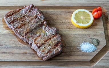 Grilled flank steak served on a wooden cutting board with lemon, cherry tomatoes, and seasoning, showcasing delicious culinary presentation