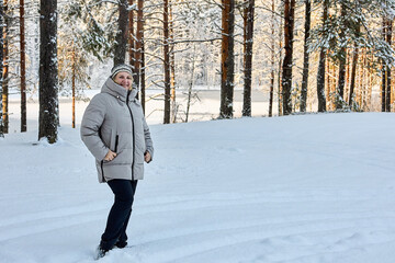 Fototapeta premium Winter walk through snowy forest, warmly dressed middle aged woman poses to someone against small frozen lake.
