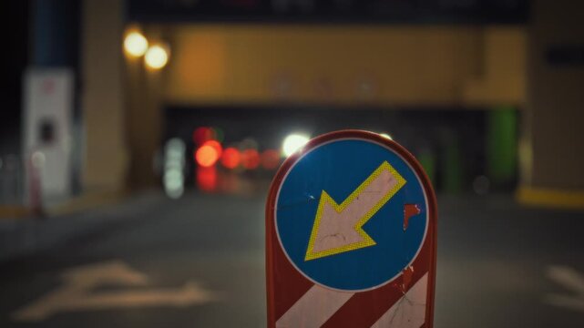 A traffic sign shows an arrow pointing left, guiding vehicles on the correct side of the road. Dim streetlights illuminate the area, while a car approaches from a distance