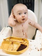 Toddler covered in food sits in a high chair and enthusiastically eating for meal. Concept of early childhood nutrition and introducing solid foods to infants.