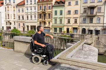 Man tourist with disability relaxes by the Ljubljana river, enjoying the European buildings.