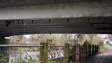mosscovered posts and calm reflections, quiet walkway beneath aged concrete bridge and wooden railings, peaceful riverside stroll under old bridge with mossy supports and reflections
