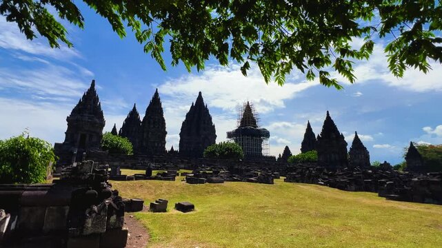 View of the ancient Prambanan Temple, a Hindu temple complex in Yogyakarta, Indonesia. The temple rises above the green foliage under a blue sky. The towers cast shadows on a sunny day. 4К