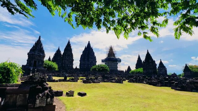 View of the ancient Prambanan Temple, a Hindu temple complex in Yogyakarta, Indonesia. The temple rises above the green foliage under a blue sky. The towers cast shadows on a sunny day. 4К
