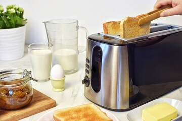 On the table is a modern toaster with slices of bread, butter, milk, and berries for breakfast. A woman removes a slice of bread from the toaster with tongs.