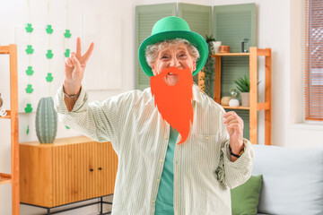 Happy senior woman in leprechaun hat with paper ginger beard showing victory gesture at home. St. Patrick's Day celebration