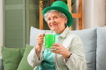 Senior woman in leprechaun hat with mug of green beer sitting on sofa at home. St. Patrick's Day celebration