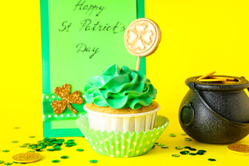 Tasty cupcake with pot of golden coins and festive postcard for St. Patrick's Day on yellow background