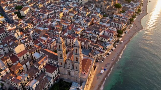 The coastal town features charming buildings with terracotta roofs lining the sunny shoreline, where waves gently lap against the sandy beach. Towering church steeples oversee the