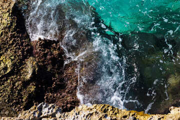 Top view of turquoise transparent wave with white foam hitting rocky seashore on a bright sunny day