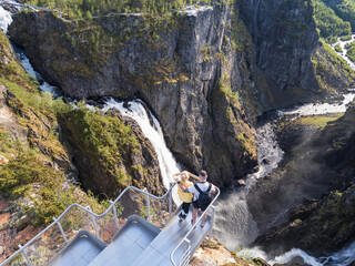 High-angle drone view of a couple on a modern viewing platform overlooking the majestic Voringsfossen waterfall in Norway.