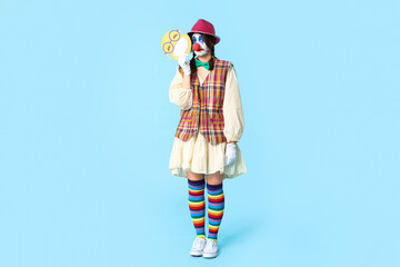Young woman dressed as sad clown with paper smile on blue background. April Fool's day celebration