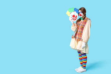 Young woman dressed as sad clown with paper joker on blue background. April Fool's day celebration