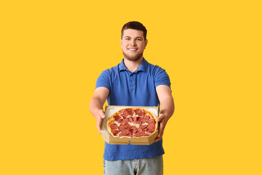 Happy young man holding cardboard box with tasty pepperoni pizza on yellow background