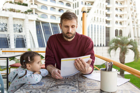 A father and his young daughter sit together at an outdoor cafe, reading the menu and choosing what to order while enjoying the warm weather and relaxed atmosphere.