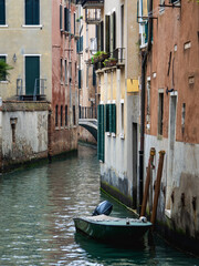 A picturesque and tranquil view of a canal (rio) in Venice, Italy. A small green motorboat is moored alongside colorful historic buildings that line the waterway.