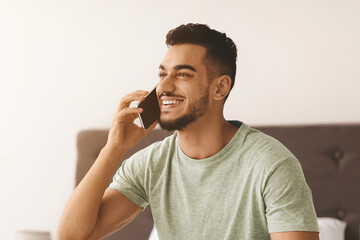 A young man with a joyful expression talks on his smartphone while sitting in a comfortable bedroom. The soft light of morning creates a warm atmosphere as he engages in conversation.