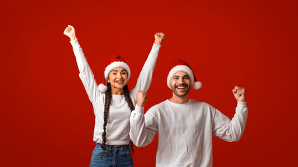 Two individuals wearing Santa hats express joy and excitement for Christmas. They are celebrating together against a vibrant red background, sharing a fun, festive moment.