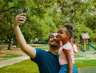 Father taking a cheerful selfie with his daughter at the park, capturing a joyful family moment outdoors. Bright colors, natural expressions and happy childhood lifestyle in a sunny environment.
