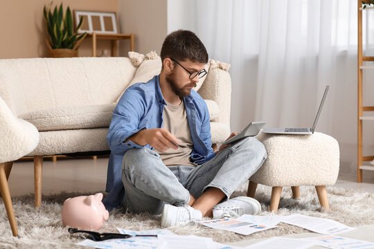 Young man in debt with calculator sitting on floor at home