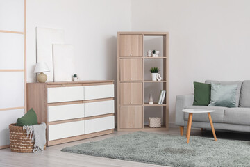 Chest of drawers with lamp and pictures, shelving unit and cozy sofa in interior of living room