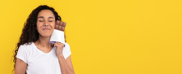 A woman with curly hair smiles blissfully while holding a chocolate bar close to her face. The...