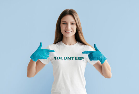 A smiling young woman in a white t-shirt with the word volunteer gestures enthusiastically with blue gloves. She is encouraging others to engage in volunteer activities for community support.
