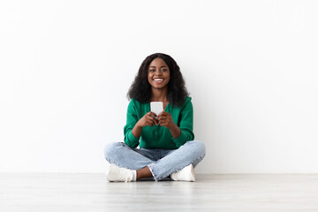 A woman sits cross-legged on the floor, wearing a green sweater and jeans. She smiles while looking at her smartphone, enjoying a moment of relaxation in a well-lit area.