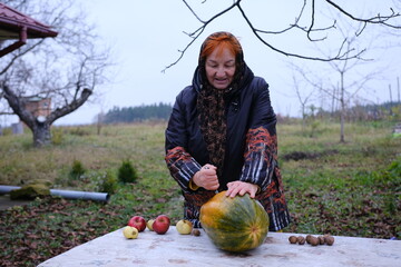 An adult red-haired woman in a garden outside on a table cutting a large pumpkin in autumn cold in...