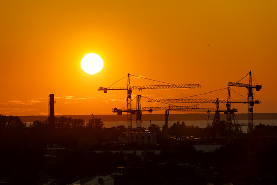 Silhouettes of multiple construction cranes and industrial structures are set against a vibrant orange sunset sky over Tallinn, with the sea visible in the distance