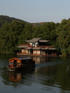 View of a traditional wooden boat glides across the tranquil lake towards a serene pavilion nestled amidst lush greenery, Hangzhou, Zhejiang, China.
