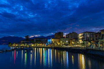 Naklejka premium Night view of the Porto Valtravaglia waterfront, featuring the brightly illuminated buildings reflecting on the calm lake surface beneath a dark, cloudy blue sky