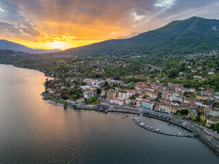 Aerial view of the coastal town of Porto Valtravaglia on Lake Maggiore at sunrise, showing the harbor, colorful buildings, and steep forested hills behind the town