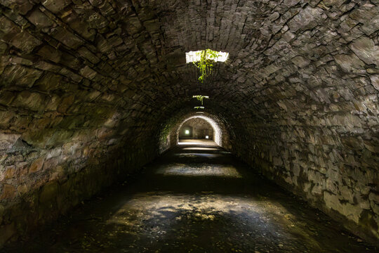 Interior view of one of the dark, vaulted stone tunnels or cisterns beneath the Castello di Brescia on Cidneo Hill, with light filtering through an overhead opening and illuminating the far end