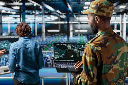 Black IT military expert in server room supervising high tech defense systems on laptop screen. Armed forces male officer doing live drone tracking, command center mission for safety.