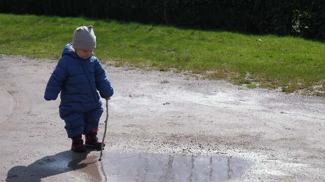 Child, dressed in cozy blue snowsuit, happily explores shimmering puddle with stick. Joyful little baby embodying the essence of playful childhood, wonder and fun in the great outdoor fun - Powered by Adobe